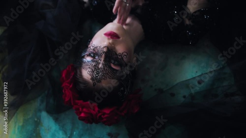 Beautiful young woman in a black masquerade mask and black peignoir lies on multi-colored floor