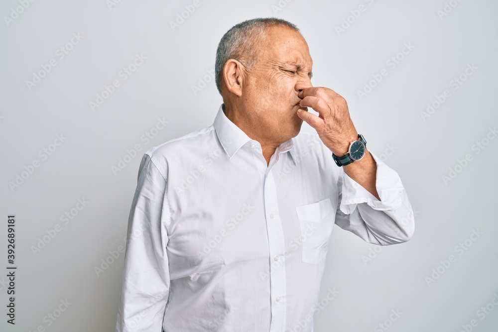Handsome senior man wearing casual white shirt smelling something stinky and disgusting, intolerable smell, holding breath with fingers on nose. bad smell Handsome senior man wearing casual white shirt smelling something stinky and disgusting, intolerable smell, holding breath with fingers on nose. bad smell