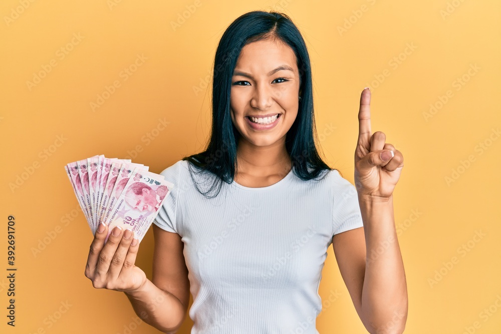 Beautiful hispanic woman holding 10 turkish lira banknotes smiling with an idea or question pointing finger with happy face, number one Beautiful hispanic woman holding 10 turkish lira banknotes smiling with an idea or question pointing finger with happy face, number one