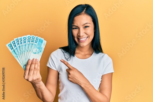 Beautiful hispanic woman holding 100 brazilian real banknotes smiling happy pointing with hand and finger photo