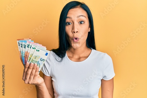 Beautiful hispanic woman holding euro banknotes scared and amazed with open mouth for surprise, disbelief face