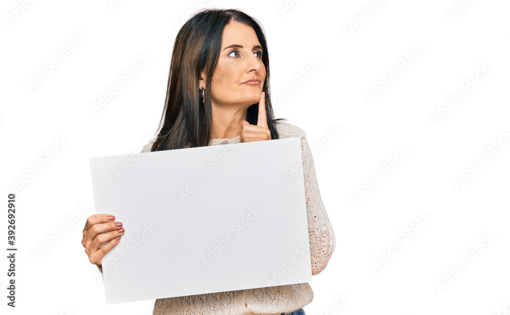 Middle age brunette woman holding blank empty banner serious face thinking about question with hand on chin, thoughtful about confusing idea Middle age brunette woman holding blank empty banner serious face thinking about question with hand on chin, thoughtful about confusing idea