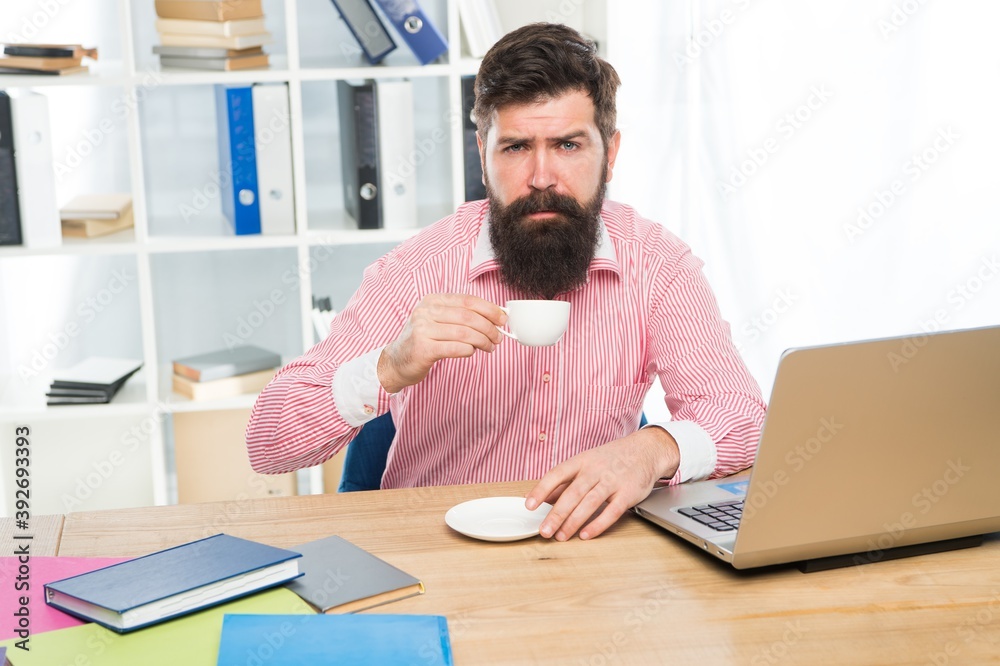 Exhausted clerical worker with hipster beard drink coffee sitting at desktop in modern office, morning