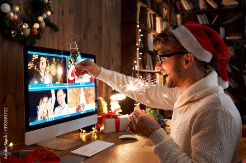 Photography Young man wearing Christmas hat drinking champagne holding sparkler talking to friends on virtual video call celebrate Happy New Year party in distance online conference chat on pc computer at home