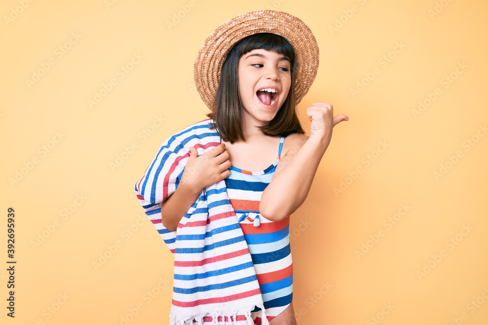 Young little girl with bang wearing swimsuit and summer hat holding towel pointing thumb up to the side smiling happy with open mouth Young little girl with bang wearing swimsuit and summer hat holding towel pointing thumb up to the side smiling happy with open mouth