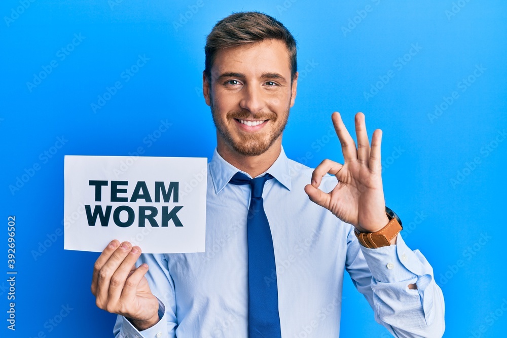 Handsome caucasian business man holding team work banner doing ok sign with fingers, smiling friendly gesturing excellent symbol Handsome caucasian business man holding team work banner doing ok sign with fingers, smiling friendly gesturing excellent symbol