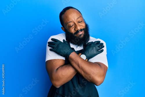 Young african american man tattoo artist wearing professional uniform and gloves hugging oneself happy and positive, smiling confident. self love and self care