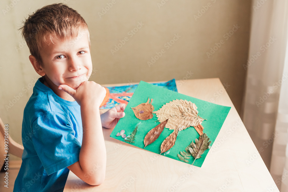 Cute boy demonstrate applique made of autumn leaves, happy child sitting by desk Cute boy demonstrate applique made of autumn leaves, happy child sitting by desk