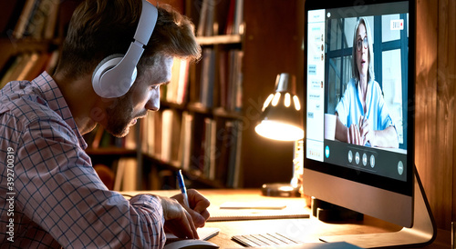 Male student wearing headphones conference video calling, watching webinar, online training class, virtual chat meeting with remote teacher or coach distance learning using computer, taking notes. photo