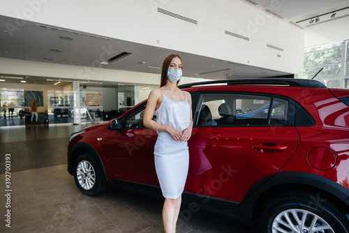 A young pretty girl inspects a new car at a car dealership in a mask during the pandemic. The sale and purchase of cars, in the period of pandemia © Andrii