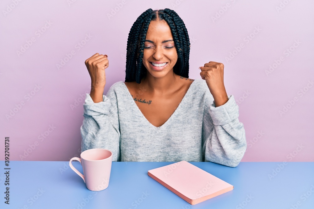 Beautiful hispanic woman reading a book and drinking a coffee very happy and excited doing winner gesture with arms raised, smiling and screaming for success. celebration concept. Beautiful hispanic woman reading a book and drinking a coffee very happy and excited doing winner gesture with arms raised, smiling and screaming for success. celebration concept.