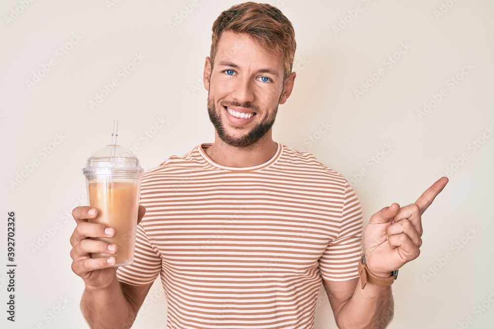 Young caucasian man drinking glass of orange juice smiling happy pointing with hand and finger to the side Young caucasian man drinking glass of orange juice smiling happy pointing with hand and finger to the side