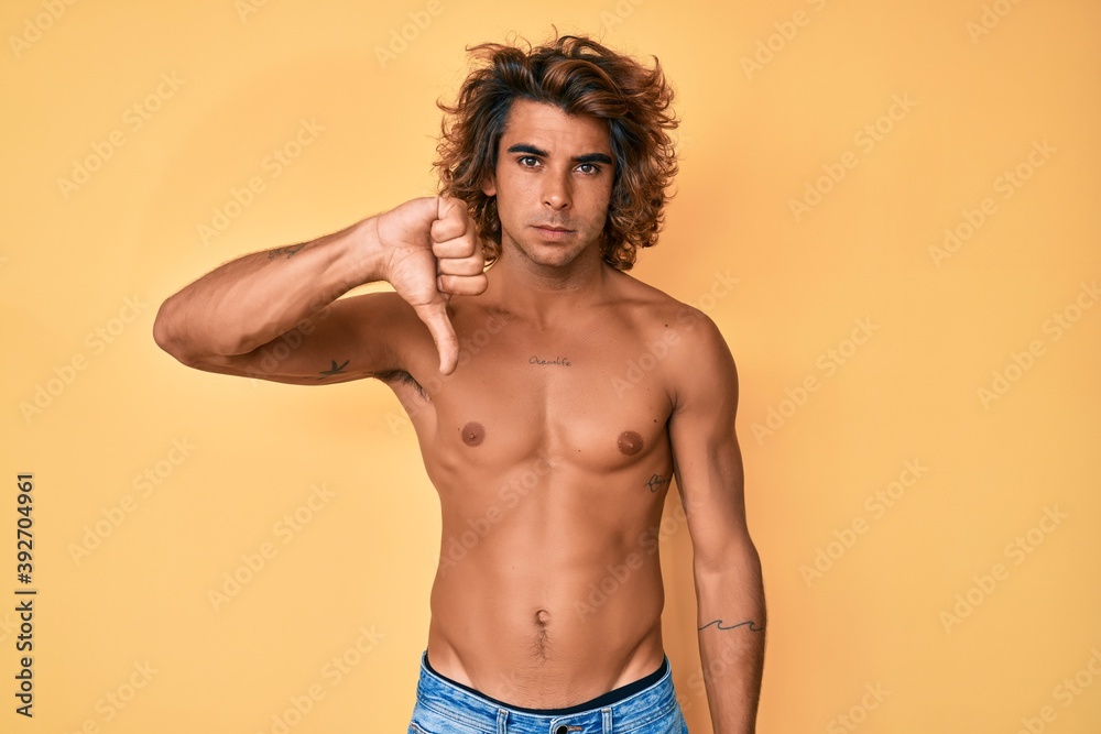 Fototapeta premium Young hispanic man standing shirtless looking unhappy and angry showing rejection and negative with thumbs down gesture. bad expression.