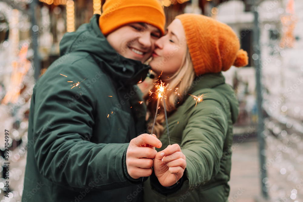 Young attractive couple hug and kiss outdoors with the sparklers in their hands celebrating New Year and Christmas. Winter holidays festival mood. Man and woman feel happy. Love and happiness concept.