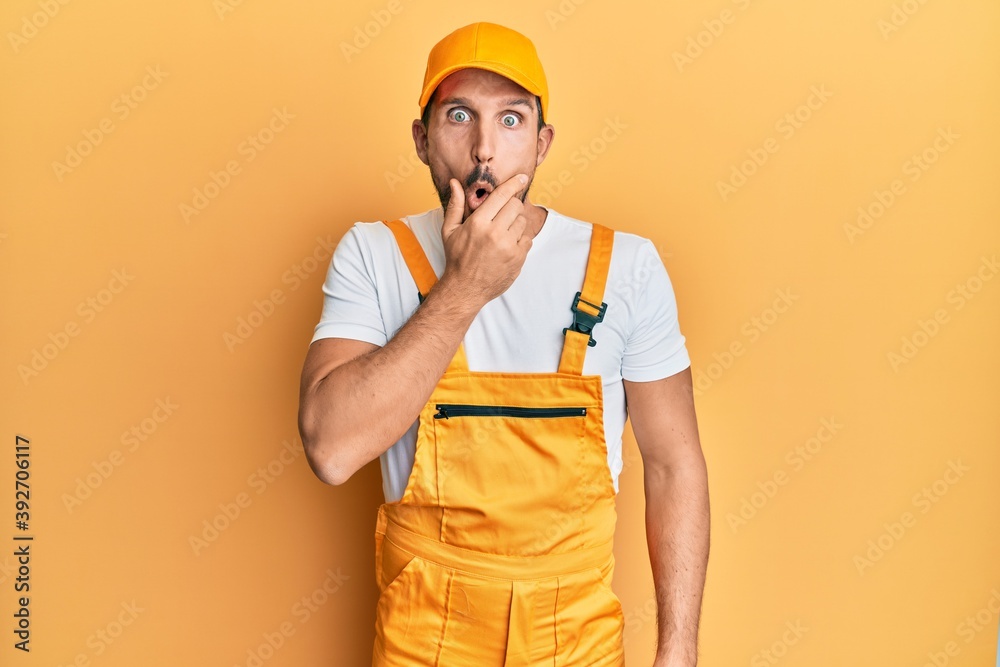 Young handsome man wearing handyman uniform over yellow background looking fascinated with disbelief, surprise and amazed expression with hands on chin