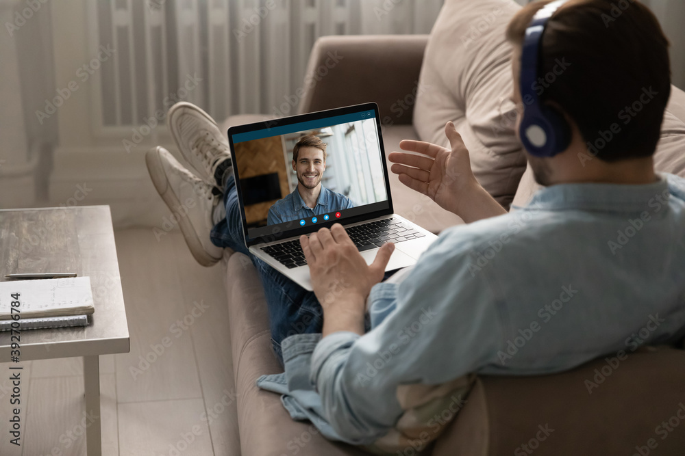 Rear back view relaxed young guy wearing headphones lying on cozy couch, enjoying fun talk with male buddy, sharing life news online using laptop video call application, distant communication concept.