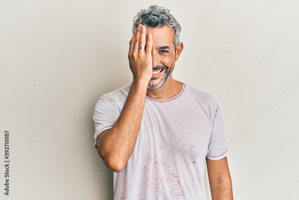 Middle age grey-haired man wearing casual clothes covering one eye with hand, confident smile on face and surprise emotion. Middle age grey-haired man wearing casual clothes covering one eye with hand, confident smile on face and surprise emotion.