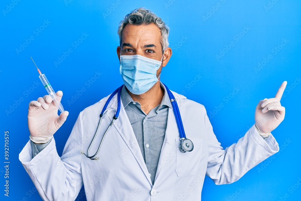Middle age grey-haired man wearing doctor uniform and medical mask holding syringe smiling happy pointing with hand and finger to the side Middle age grey-haired man wearing doctor uniform and medical mask holding syringe smiling happy pointing with hand and finger to the side