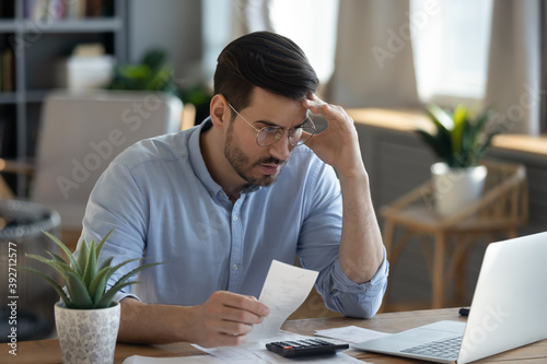 Small business owner experiences financial crisis, high costs, bank debt, money overspend concept. Man sitting at desk looks at laptop screen holding receipt feels desperate after calculating expenses photo