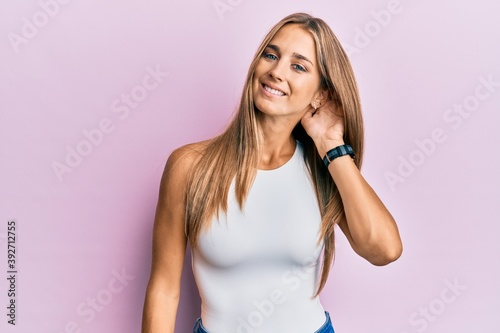 Young blonde woman wearing casual clothes smiling with hand over ear listening and hearing to rumor or gossip. deafness concept.