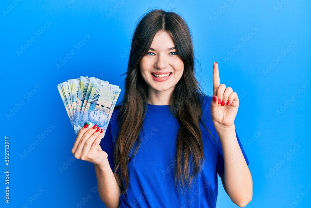 Young beautiful caucasian girl holding south african 100 rand banknotes smiling with an idea or question pointing finger with happy face, number one Young beautiful caucasian girl holding south african 100 rand banknotes smiling with an idea or question pointing finger with happy face, number one