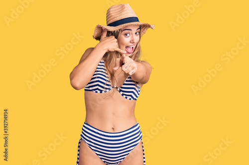 Young beautiful blonde woman wearing bikini and hat smiling doing talking on the telephone gesture and pointing to you. call me.