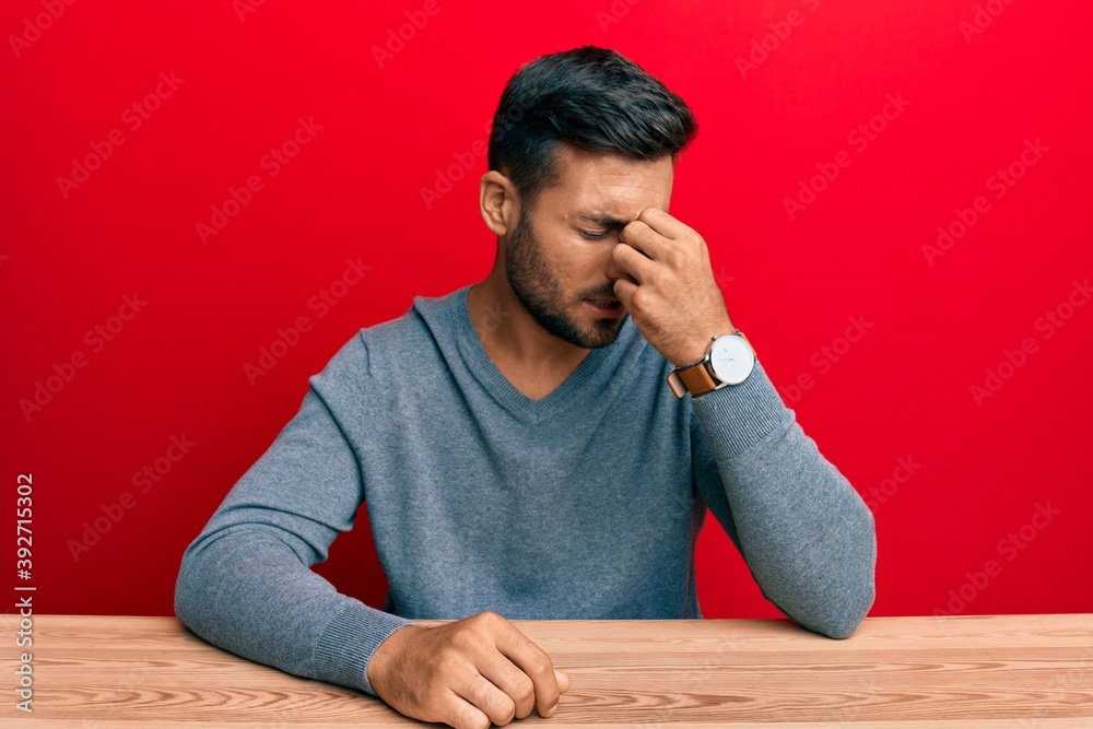 Handsome hispanic man wearing casual style sitting on the table tired rubbing nose and eyes feeling fatigue and headache. stress and frustration concept. Handsome hispanic man wearing casual style sitting on the table tired rubbing nose and eyes feeling fatigue and headache. stress and frustration concept.