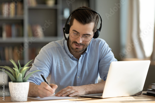 Focused 35s man sit at desk wear headphones watch webinar use laptop gain new knowledge writing notes. Video conference communication, negotiation remotely, study online, e-learn, self-educate concept photo