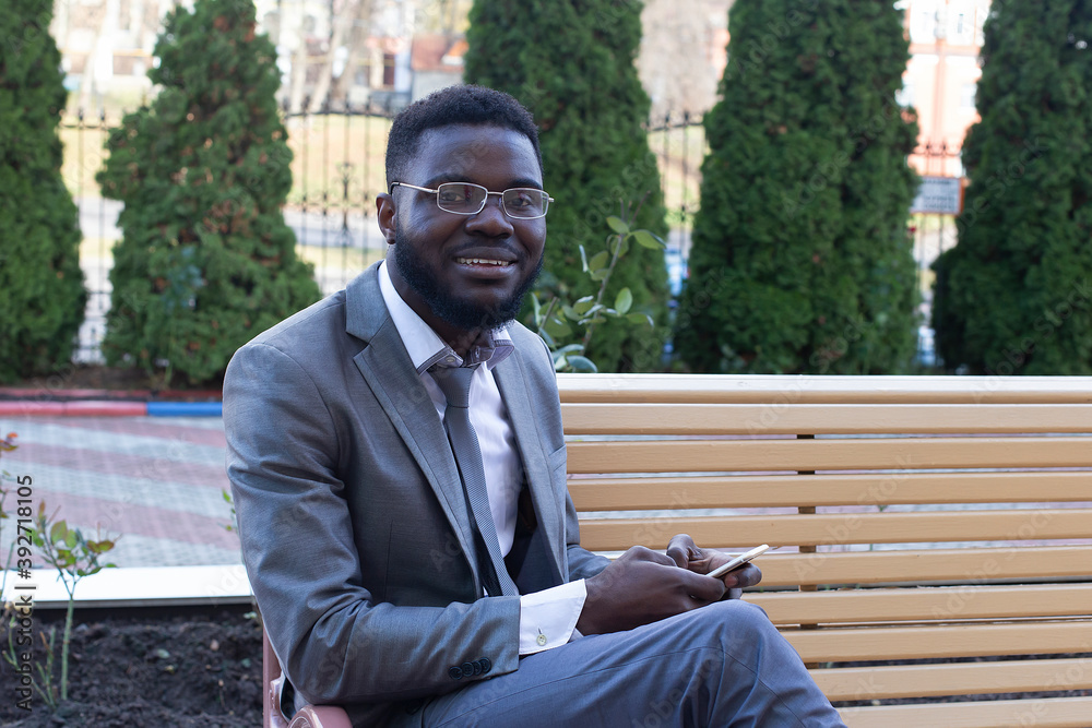 Fototapeta premium Young African businessman smiling on city bench with phone, tablet