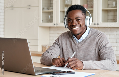 Happy african business man, black male student wearing headphones elearning on laptop sitting at kitchen table working from home office, learning online, studying remote course looking at camera.
