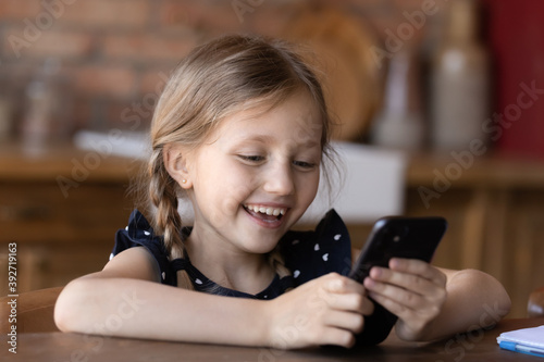 Kid and gadget. Happy laughing little child girl sitting at desk holding mobile phone in hands, making call to mother, chatting with friend, watching funny cartoon, posting selfie in social network