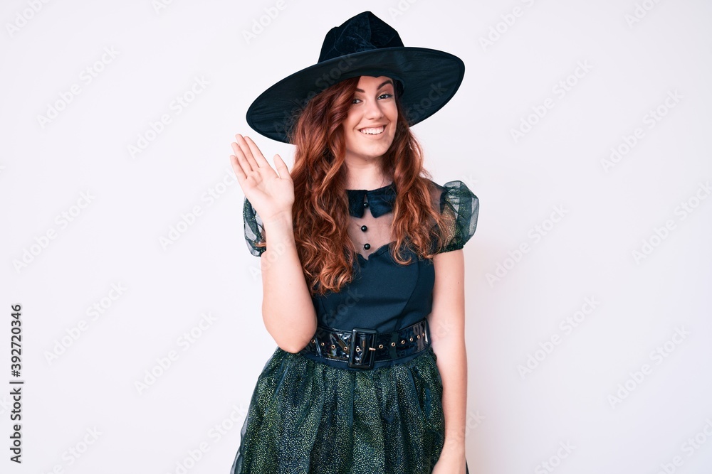 Young beautiful woman wearing witch halloween costume waiving saying hello happy and smiling, friendly welcome gesture Young beautiful woman wearing witch halloween costume waiving saying hello happy and smiling, friendly welcome gesture