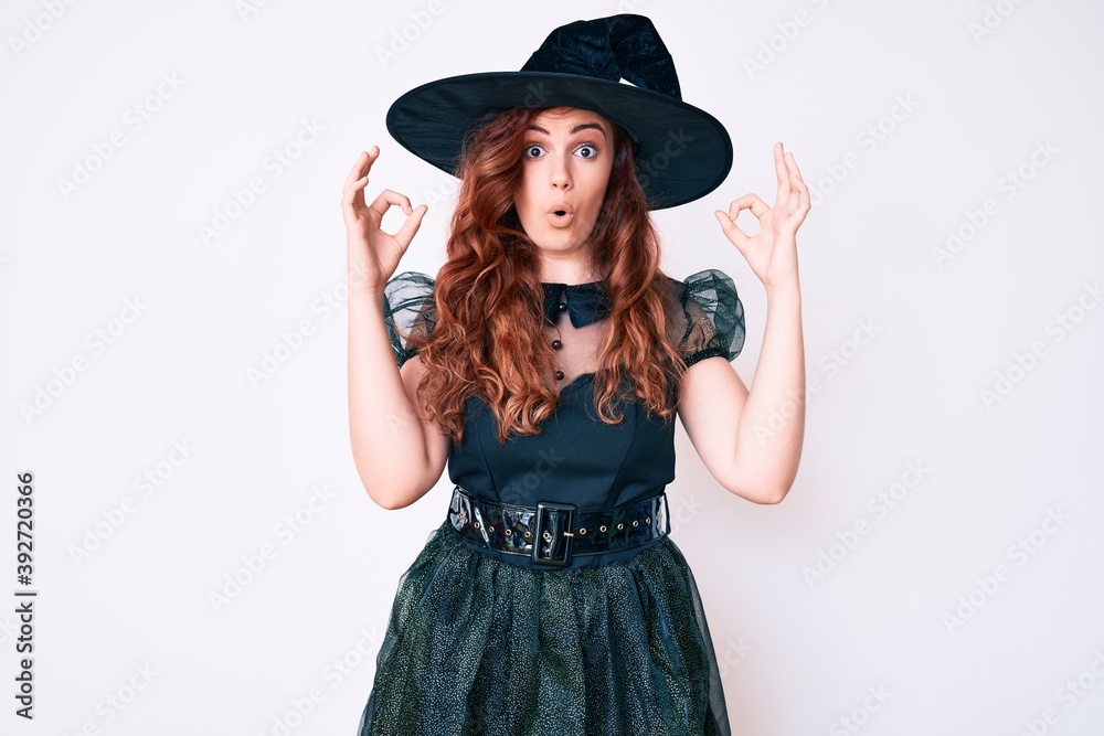 Young beautiful woman wearing witch halloween costume looking surprised and shocked doing ok approval symbol with fingers. crazy expression Young beautiful woman wearing witch halloween costume looking surprised and shocked doing ok approval symbol with fingers. crazy expression