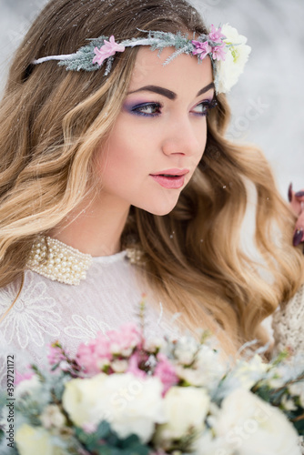 Cute bride in the winter snow forest outdoors with beautiful flowers on her head and a bouquet. Wedding makeup.