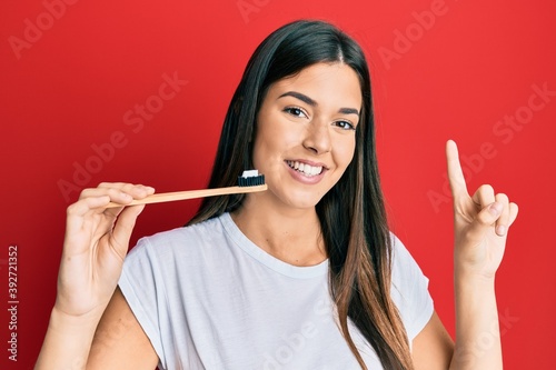 Fototapeta Naklejka Na Ścianę i Meble -  Young brunette woman holding toothbrush with toothpaste smiling with an idea or question pointing finger with happy face, number one
