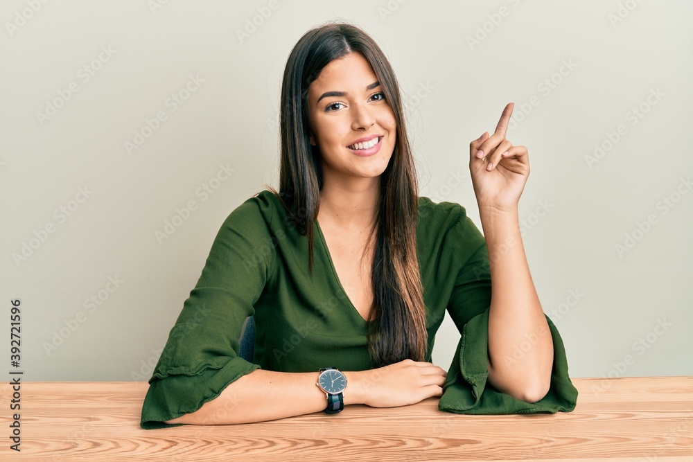 Young brunette woman wearing casual clothes sitting on the table with a big smile on face, pointing with hand and finger to the side looking at the camera. Young brunette woman wearing casual clothes sitting on the table with a big smile on face, pointing with hand and finger to the side looking at the camera.