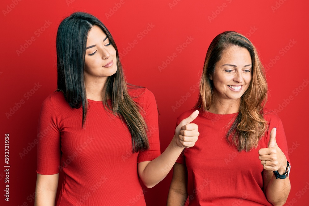 Hispanic family of mother and daughter wearing casual clothes over red background looking proud, smiling doing thumbs up gesture to the side