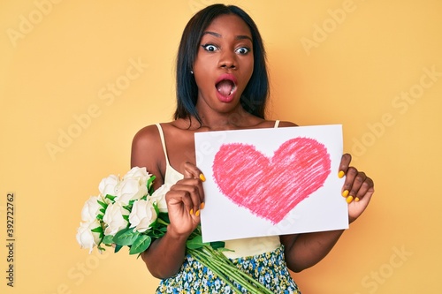 Young african american woman holding heart draw and flowers afraid and shocked with surprise and amazed expression  fear and excited face.