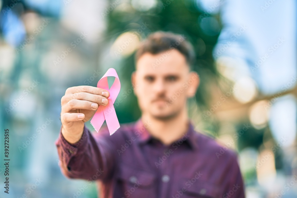 Young hispanic man with serious expression holding pink breast cancer ribbon at the city.