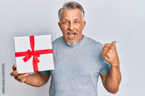 Middle age grey-haired man holding gift pointing thumb up to the side smiling happy with open mouth