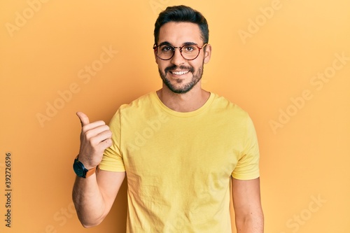 Young hispanic man wearing casual clothes and glasses smiling with happy face looking and pointing to the side with thumb up.