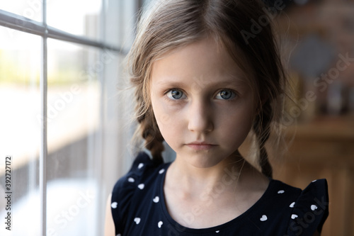 Fotografía Close up portrait of pensive serious little school preschool age girl standing by window at home looking at camera with sad unhappy face having problem trouble thinking about bad relations in family