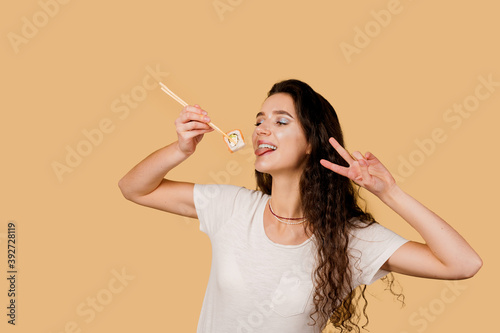 Happy girl with sushi isolated on yellow background. Young girl is holding sushi by chopsticks, smiling, having fun and wanting to eat one piece of roll philadelphia. Advert for sushi delivery.