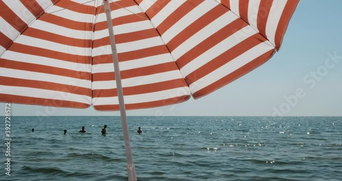 Sunny summer day with horizontal shot of a sea, ocean, sand with people swimming and red striped umbrella creating shadow. Water with shiny reflections. Vacation, holiday, island chill relaxation mood