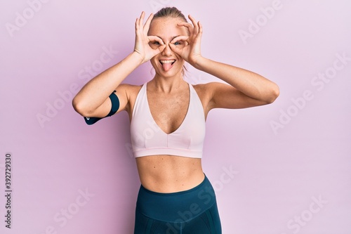 Beautiful blonde woman wearing sportswear and arm band doing ok gesture like binoculars sticking tongue out, eyes looking through fingers. crazy expression. © Krakenimages.com