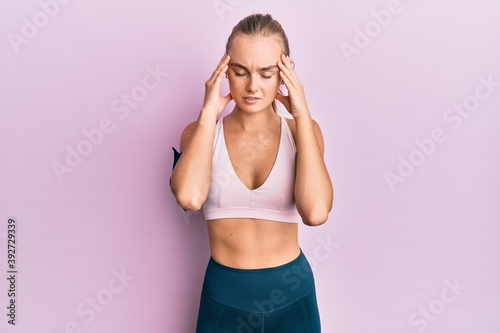 Beautiful blonde woman wearing sportswear and arm band with hand on head for pain in head because stress. suffering migraine.