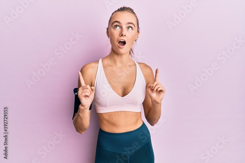Beautiful blonde woman wearing sportswear and arm band amazed and surprised looking up and pointing with fingers and raised arms. © Krakenimages.com