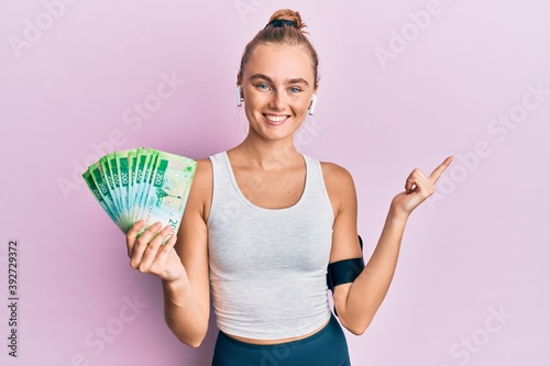 Beautiful blonde sport woman holding russian 100 ruble banknotes smiling happy pointing with hand and finger to the side