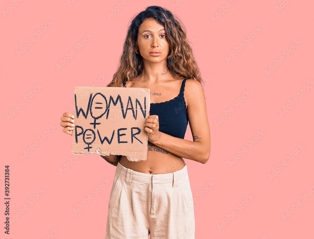 Young hispanic woman with tattoo holding woman power banner thinking attitude and sober expression looking self confident Young hispanic woman with tattoo holding woman power banner thinking attitude and sober expression looking self confident