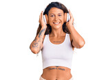 Young hispanic woman with tattoo listening to music using headphones looking positive and happy standing and smiling with a confident smile showing teeth